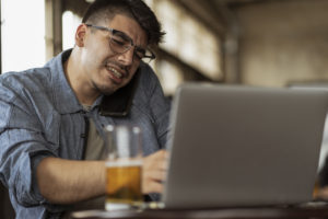 Man working on laptop and talking on phone with beer glass on table